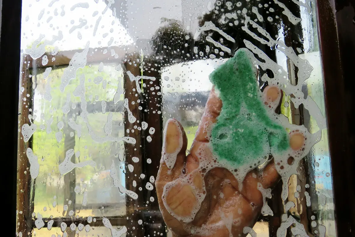 A hand pressing a sponge to wet window glass with soap bubbles and streaks