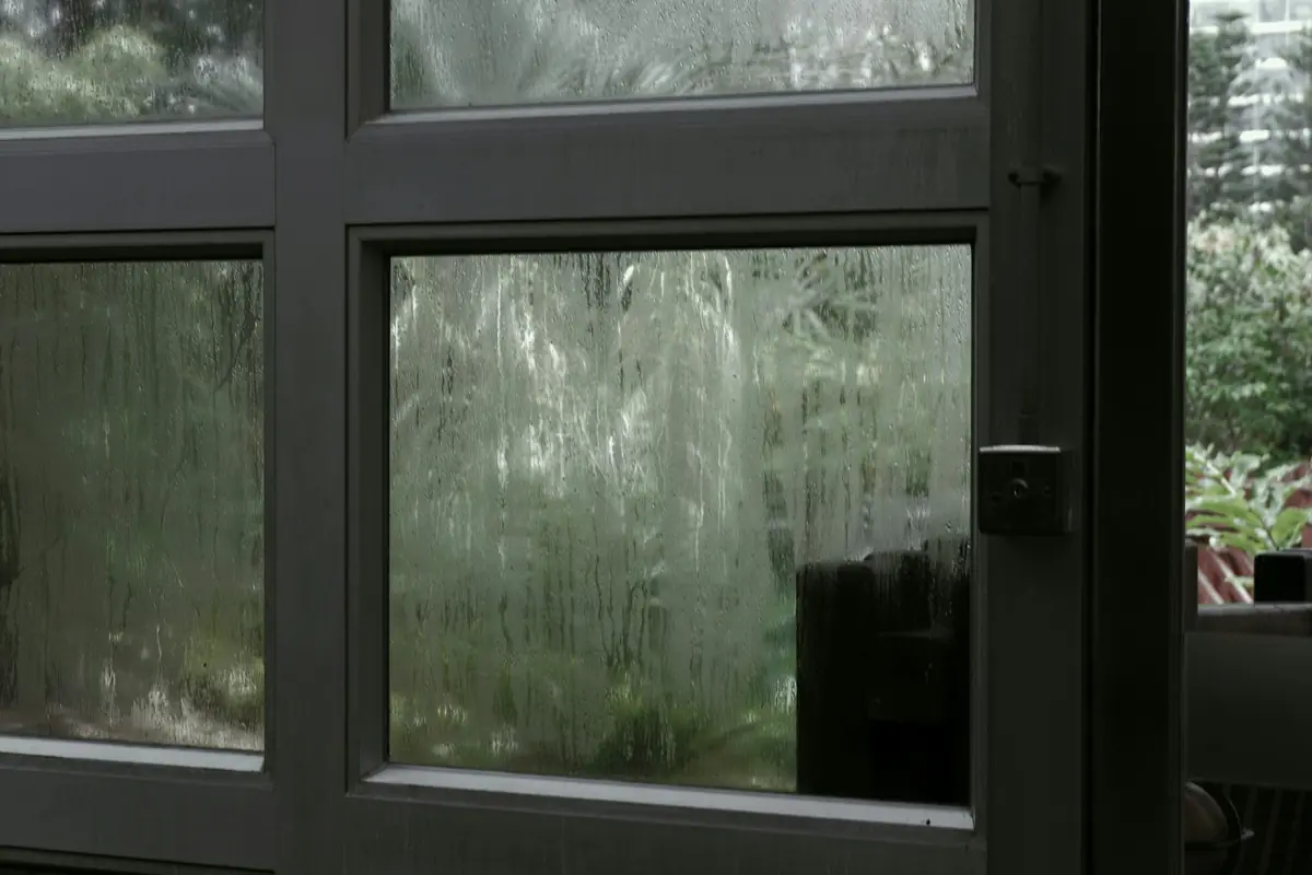 Water streaking down the panes of a black-framed window with greenery and a tower outside