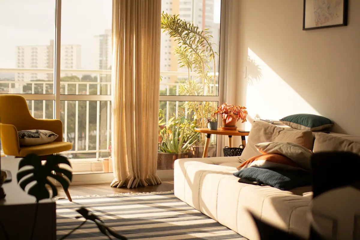Living room in warm morning light with a sofa, sheer curtain and a plant-filled balcony beyond
