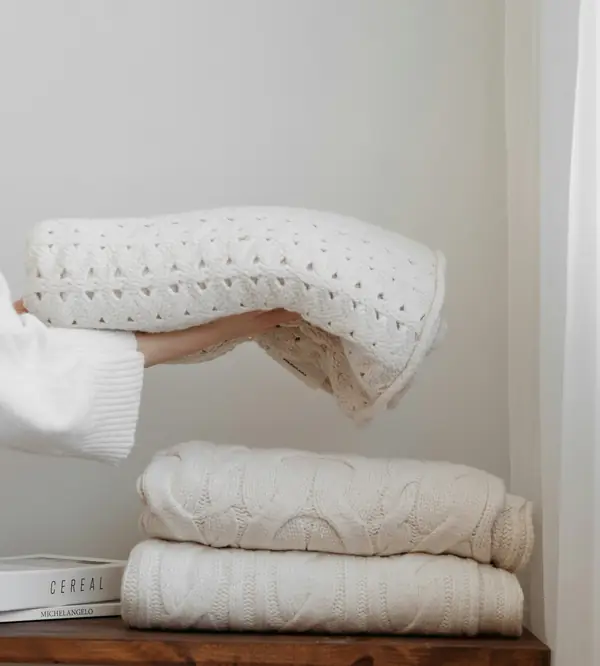 Hands placing a folded cream knit blanket on a neat stack of throws
