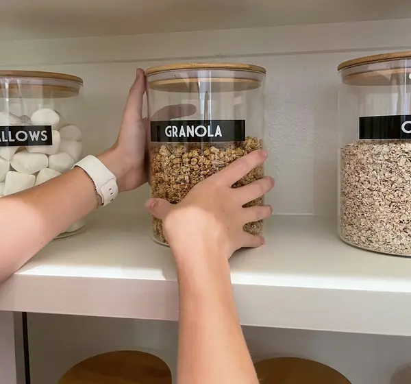 Hands placing a labelled glass jar on a neat white pantry shelf