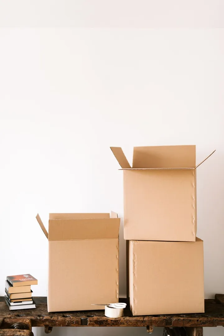 Cardboard moving boxes and a roll of packing tape on a wooden bench against a white wall