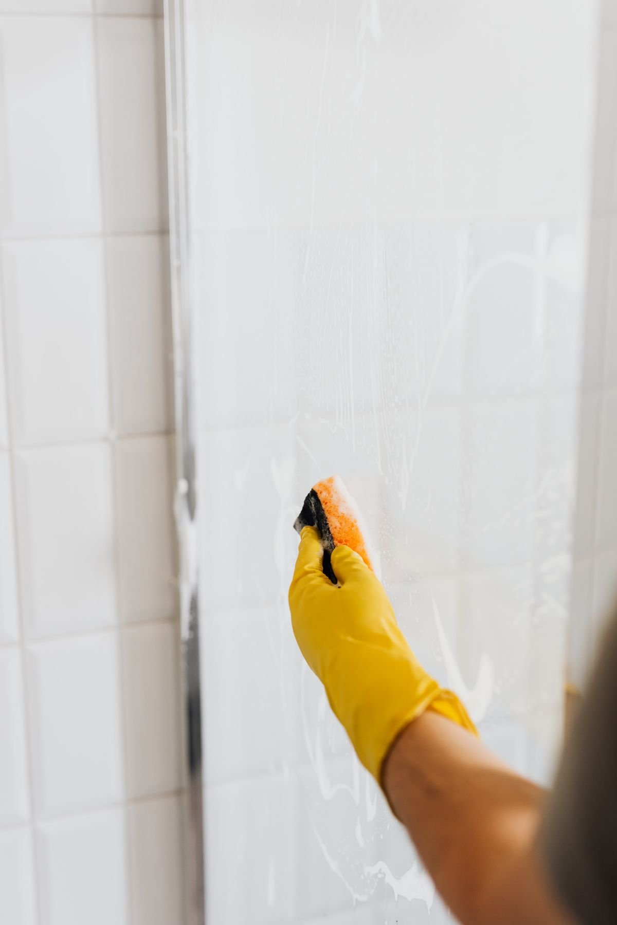 Hand in yellow glove scrubbing a soapy shower door with a sponge
